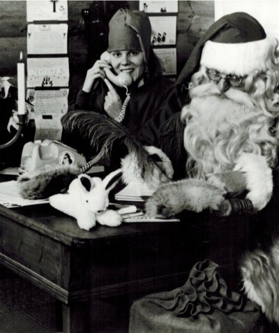 santa sits at a desk writing his list while an elf answers a phone next to him